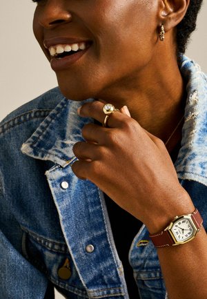 Montre avec un bracelet en cuir marron, un boîtier octogonal en or, un cadran blanc et des chiffres romains, portée avec une veste en denim et une bague en or.