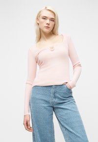 Ξανθιά γυναίκα που φοράει ανοιχτό ροζ μακρυμάνικο τοπ με μοναδικό ντεκολτέ και μπλε παντελόνι με φαρδιά πόδια και σχέδιο, στέκεται με το ένα χέρι στην τσέπη.