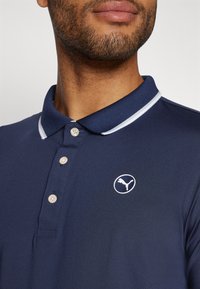 Polo navy en tissu lisse avec un bord blanc au col. Il dispose d'une patte de boutonnage à trois boutons et d'un logo Puma blanc sur la poitrine.