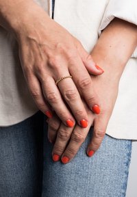 Inel cu mărgele aurii pe mâini cu ojă portocalie. Pe deasupra se vede un top din țesătură de culoare deschisă și blugi albaștri. Mâinile sunt ușor împletite.