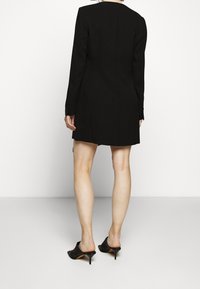 Robe blazer noire taillée sur mesure avec manches longues, coupe ajustée, texture lisse et détails subtils de coutures, associée à des mules à talons noires.