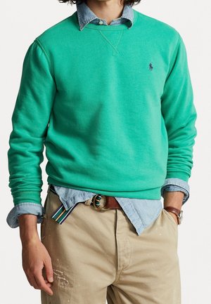 Homme portant un sweat-shirt bleu sarcelle par-dessus une chemise bleu clair à boutons, pantalon beige avec ceinture marron, et montre-bracelet marron, les mains dans les poches.