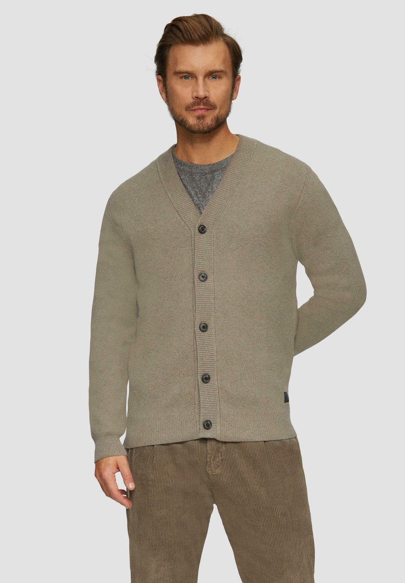 Khakifarbener Cardigan mit V-Ausschnitt, gerippten Bündchen und Saum, ausgestattet mit sechs schwarzen Knöpfen. Getragen über einem grauen T-Shirt, mit strukturierten Cordhosen.