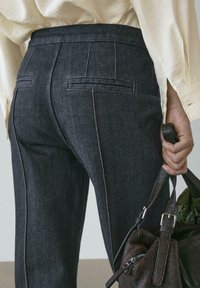 Dunkle Denimhose mit tailliertem Schnitt, auffälligen Nähten, zwei Gesäßtaschen und einer glatten Textur. Eine dunkle Handtasche wird gehalten.