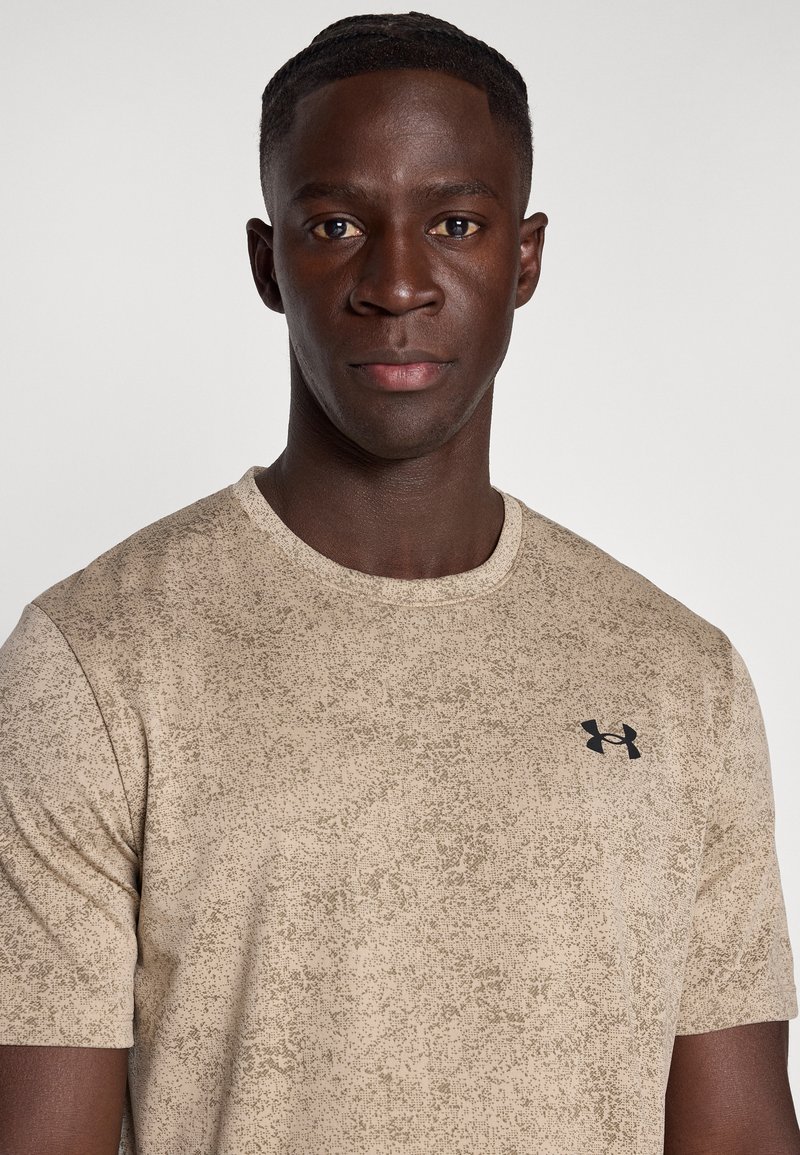 Joven con el pelo corto que lleva una camiseta beige moteada de Under Armour, mirando hacia adelante con una expresión neutral sobre un fondo liso.