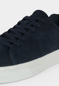 Gros plan d'une basket en daim bleu foncé avec des lacets assortis et une semelle en caoutchouc blanche sur un fond uni.