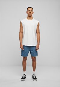 T-shirt sans manches blanc, shorts en denim, baskets noires avec semelles blanches et chaussettes blanches. Tenue décontractée avec une coupe ample.