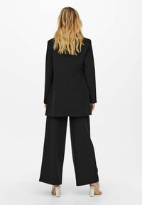 Blondi nainen seisoo selkä kameraan, päällä pitkä musta bleiseri, leveät mustat housut ja läpinäkyvät korkokengät valkoista taustaa vasten.