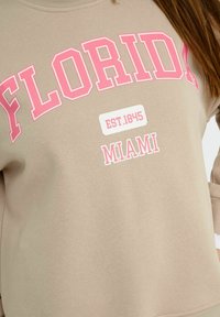 Sweatshirt beige avec un texte rose vif : "FLORIDA", "EST. 1845" et "MIAMI". Tissu doux avec un col rond.
