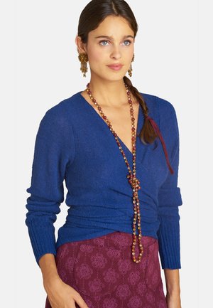 Frau mit geflochtenem Haar, die einen blauen Wickelpullover, eine lange Perlenkette, goldene Ohrringe und einen bordeauxfarbenen gemusterten Rock trägt.