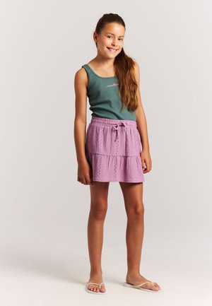 Grünes Tanktop mit pinken Buchstaben kombiniert mit einem mehrstufigen, hellvioletten Rock mit Lochstickereien. Model trägt leichte Sandalen.