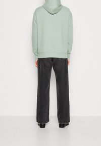 Sudadera verde clara con capucha ajustable, combinada con unos jeans oscuros de corte holgado en un gris antracita y botas negras. Los materiales parecen suaves.