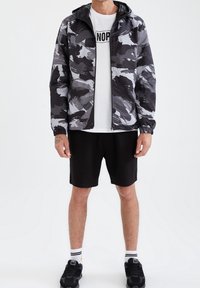 Grau-schwarze Tarnjacke mit Kapuze, strukturiertem Stoff, elastischen Bündchen, getragen über einem weißen T-Shirt, kombiniert mit schwarzen Shorts und Turnschuhen.