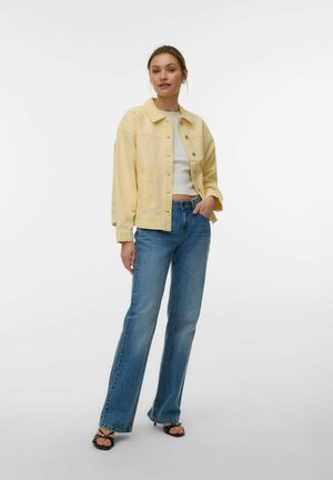 Kvinde står iført en lysegul jakke med knapper, hvid cropped top, blå jeans og sorte åbne hæle mod en ensfarvet hvid baggrund.