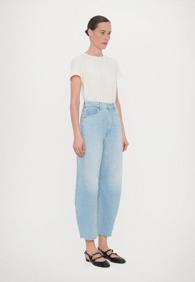 High-waisted light blue denim jeans, relaxed fit, with a plain white short-sleeve top and black strap shoes, standing on a plain background.