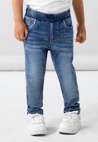 Pantalon de jogging en denim bleu avec taille élastique et cordon de serrage, présentant des poches avant et une coupe slim. Porté avec des baskets blanches.