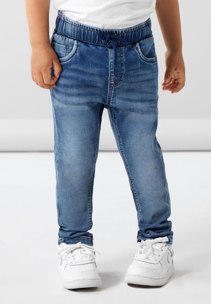 Pantalon de jogging en denim bleu avec taille élastique et cordon de serrage, présentant des poches avant et une coupe slim. Porté avec des baskets blanches.