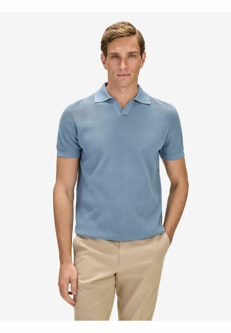 Homme aux cheveux châtain clair portant un polo en maille bleu clair à manches courtes et un pantalon beige, debout avec une main dans la poche sur un fond blanc.