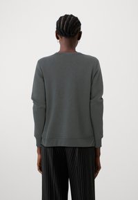 Felpa grigia con orlo e polsini a coste, texture morbida, collo a giro, indossata sopra pantaloni neri a righe, vista da dietro.