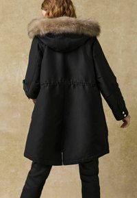 Parka noire avec une capuche bordée de fourrure, dotée d'une taille cintrée, de longues manches et de côtés zippés. Finition extérieure lisse, design de longueur genou.