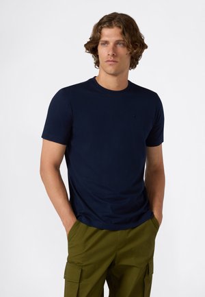 Giovane uomo che indossa una t-shirt aderente blu navy e pantaloni cargo verde oliva, in piedi con le mani in tasca contro uno sfondo bianco semplice.