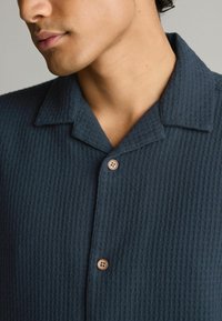 Camisa de tela texturizada azul oscura con cuello y detalles de botones marrones, mostrando una superficie acanalada y un diseño estructurado.