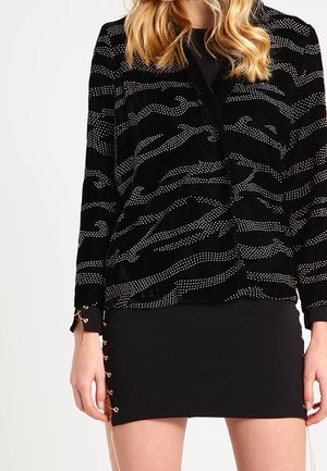 Vrouw die een zwart jasje met een patroon van witte gestippelde strepen draagt en een zwarte minirok met gouden veterdetails met oogjes.