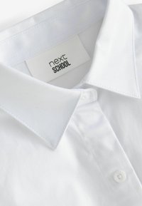 Chemise à col blanc avec une texture de tissu lisse. Comprend une patte de boutonnage et une étiquette indiquant "next SCHOOL" cousue à l'intérieur du col.