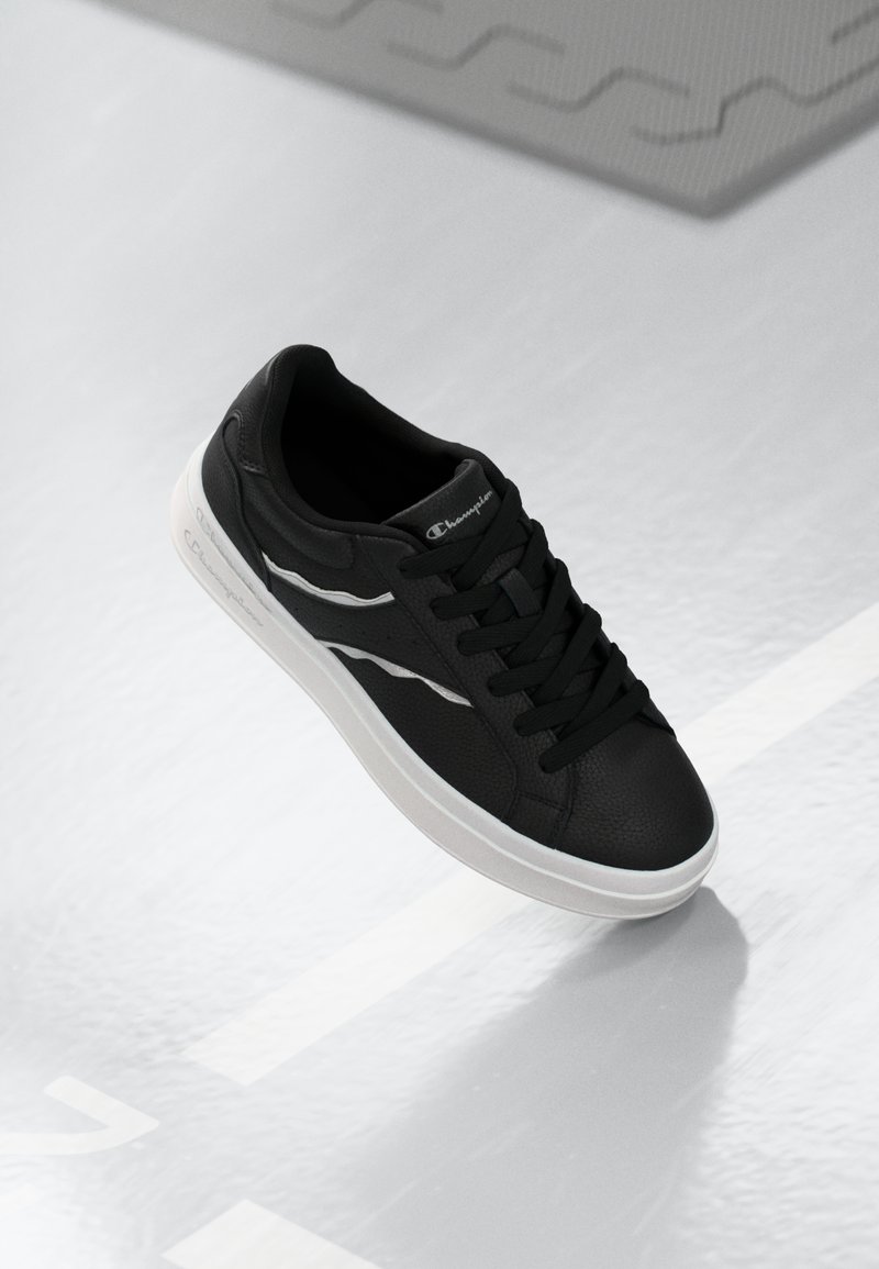Tenis negri cu un material texturat, accente albe, design cu șireturi și vârf rotund. Are un logo de marcă pe limbă.