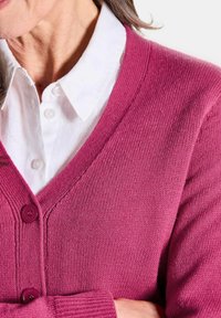 Fuchsia Cardigan mit V-Ausschnitt, strukturiertem Strickmuster und zwei lila Knöpfen. Weißes Hemd mit Kragen ist sichtbar darunter.