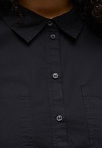 Κοντινό πλάνο για το γιακά και τα κουμπιά εμπρός σε μαύρο πουκάμισο, με ορατές ραφές και υφή υφάσματος.