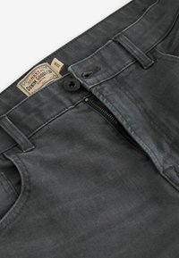 Jean slim gris foncé avec ceinture déboutonnée et fermeture éclair, montrant l'étiquette taille 32R à l'intérieur de la ceinture.