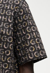 Μαύρο κοντομάνικο πουκάμισο με επαναλαμβανόμενο μοτίβο κειμένου "HUGO" σε απαλούς γκρι και κίτρινους τόνους.