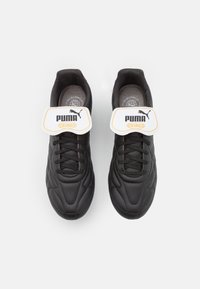 Crne sintetičke cipele s teksturiranom površinom, s bijelom trakom s natpisom "PUMA KING" u crnoj i narandžastoj boji, te crnim vezicama.