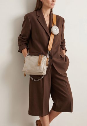 Žena v hnědém oversized blejzru a krátkých šortkách, která nosí světle béžovou kabelku přes rameno s opáleným popruhem a připevněnou malou kulatou taštičkou.