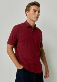 Bordo polo marškiniai iš minkštos medvilnės, su apykakle ir trumpomis rankovėmis, derinami su tamsiai džinsais. Diskretiškas logotipas ant krūtinės.