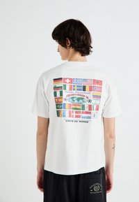 T-shirt bianca in cotone con una grafica di varie bandiere nazionali sulla schiena, con il testo "Campionato di Calcio da Tavolo" e un design del globo.