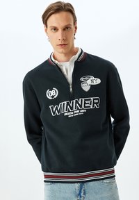 Sudadera negra con cremallera, texto y parches en blanco, cuello acanalado con rayas rojas y blancas, y tejido suave y texturizado.
