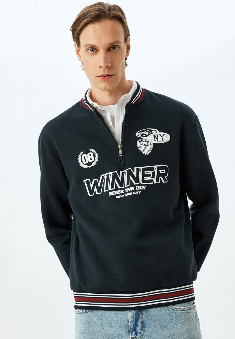 Sudadera negra con cremallera, texto y parches en blanco, cuello acanalado con rayas rojas y blancas, y tejido suave y texturizado.
