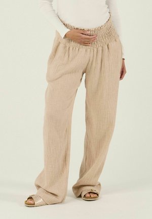 Personne portant un pantalon beige texturé ample et des sandales dorées, avec une main reposant sur leur ventre et un haut blanc à manches longues.