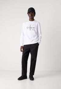 Felpa bianca con logo grigio "Calvin Klein", abbinata a pantaloni neri e sneakers nere. Il modello indossa un berretto nero.