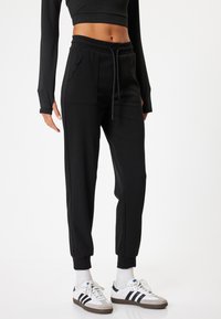 Schwarze Jogginghose mit einem Kordelzugbund, seitlichen Taschen und schmalen Bündchen, kombiniert mit weißen Sportschuhen und weißen Socken.