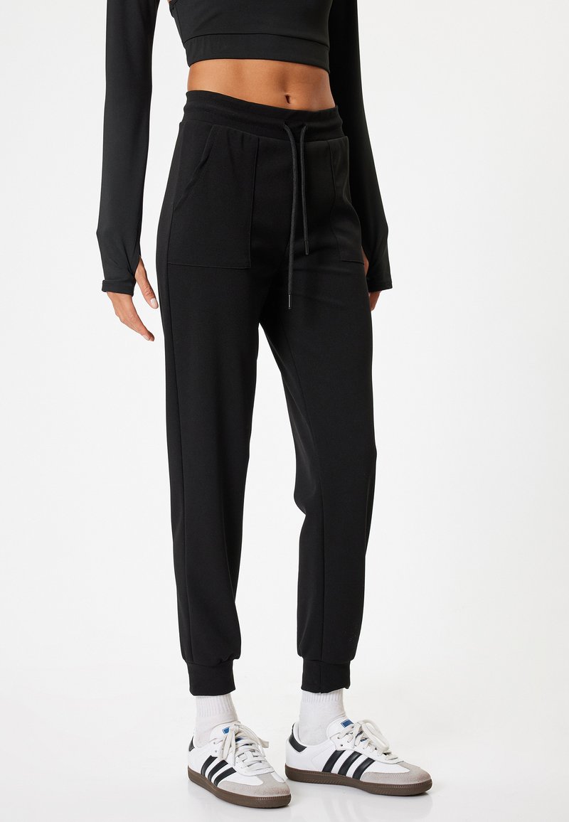 Schwarze Jogginghose mit einem Kordelzugbund, seitlichen Taschen und schmalen Bündchen, kombiniert mit weißen Sportschuhen und weißen Socken.