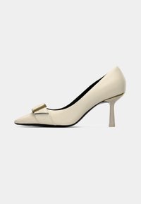 Beige high heel shoe with a pointed toe, smooth synthetic material, gold accent hardware on the front, and a slender, clear heel.