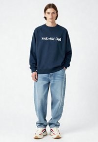 Sweat-shirt bleu marine avec le texte "YOUR ONLY FANS", associé à un jean ample bleu clair et des baskets blanches avec des accents rouges et bleus.