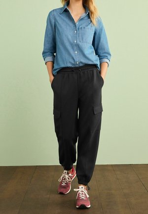 Person trägt ein blaues Hemd mit Knöpfen und hochgekrempelten Ärmeln, schwarze Cargohosen und pinke Laufschuhe und steht auf einem Holzboden.