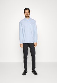 Camisa de botones azul claro con cuello, manga larga y un pequeño bolsillo en el pecho. Combinada con pantalones negros de corte slim y zapatos negros.