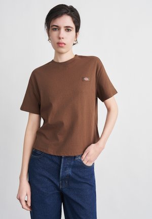 Noor lühikeste tumedate juustega, pruunis lühikeste varrukatega särgis väikese logoga ja tumesinistes teksapükstes, seisab heleda tausta ees.