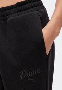 Gros plan d'un pantalon de survêtement noir Puma avec taille élastique et main dans la poche latérale.