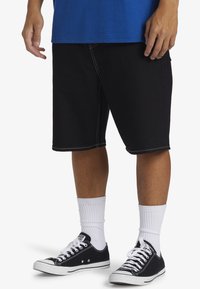 Pantalones cortos de mezclilla negros con costuras blancas en contraste, con un ajuste relajado. Combinados con zapatillas negras y calcetines blancos hasta el tobillo.